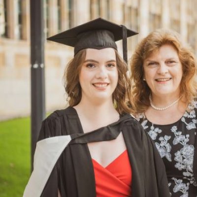 A young woman in graduation garb smiles next to an older woman in UQ's Great Court.